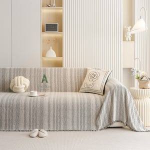 May include: A neutral-toned sofa with a patterned throw blanket and decorative pillows. The throw has a black chevron design. A white knot pillow and a pillow with a floral design are on the sofa. A pair of slippers and a teacup are on the sofa.