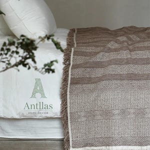 Pu&ograve; includere: Una coperta marrone intrecciata con un motivo strutturato e frange, drappeggiata su un letto con lenzuola bianche. La coperta presenta strisce orizzontali e una palette di colori neutri. Il letto ha un cuscino bianco e un copriletto bianco. Il testo "Antllas HOME DECOR" &egrave; visibile.