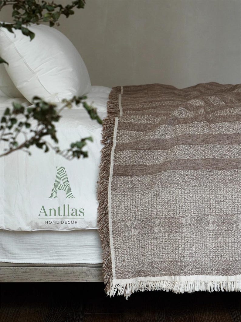 Pu&ograve; includere: Una coperta marrone intrecciata con un motivo strutturato e frange, drappeggiata su un letto con lenzuola bianche. La coperta presenta strisce orizzontali e una palette di colori neutri. Il letto ha un cuscino bianco e un copriletto bianco. Il testo "Antllas HOME DECOR" &egrave; visibile.