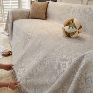 May include: A light grey sofa throw with a geometric design, fringed edges, and a matching cushion. A decorative ball-shaped cushion is also on the sofa. The sofa throw is draped over a light-coloured sofa.