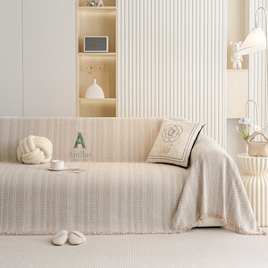 May include: Beige sofa with a patterned throw blanket and a decorative pillow featuring a rose design. A matching knot pillow, a cup, and slippers are on the sofa. The word "A" and the brand name "Antllas" are printed on the sofa.
