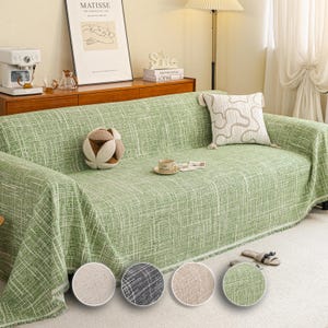 Può includere: Una fodera per divano verde con una trama intrecciata, drappeggiata su un divano. La fodera ha una frangia sul bordo inferiore. Un cuscino decorativo e un oggetto decorativo rotondo e multicolore sono sul divano. I campioni di colore sono mostrati in basso.