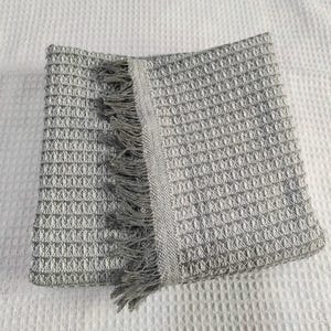 May include: A folded gray and white woven throw blanket with a textured pattern and fringe detail. The blanket is made of a soft material and is suitable for use as a decorative accent or for warmth.