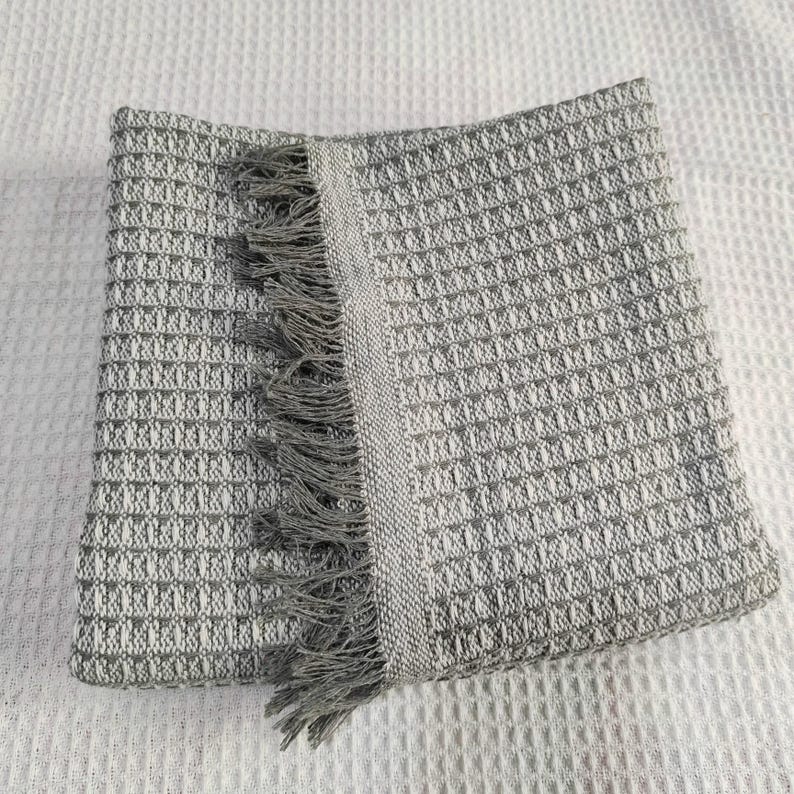 May include: A folded gray and white woven throw blanket with a textured pattern and fringe detail. The blanket is made of a soft material and is suitable for use as a decorative accent or for warmth.