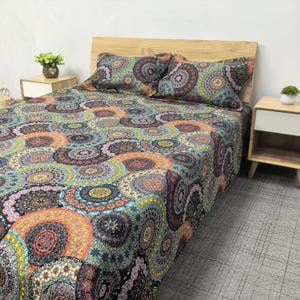 Op de afbeelding: Een bed met een patroon dekbedovertrek en twee bijpassende kussenslopen. Het ontwerp heeft een herhalend cirkelvormig patroon in tinten teal, oranje, bruin en geel. Het hoofdbord is gemaakt van lichtgekleurd hout.