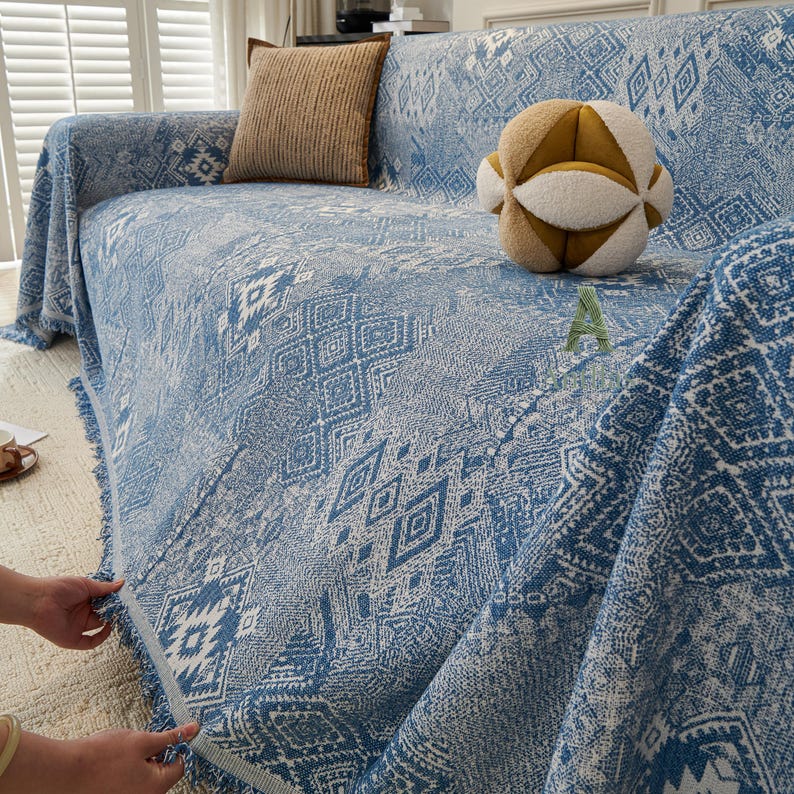 May include: A blue and white patterned sofa cover with a geometric design. The cover is draped over a sofa, with a fringed edge. A decorative cushion and a spherical plush toy are on the sofa.