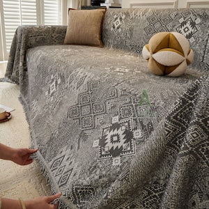 May include: A grey sofa cover with a geometric pattern and fringed trim. The cover is draped over a sofa, with a brown cushion and a decorative ball on top. The pattern includes white and dark grey shapes.