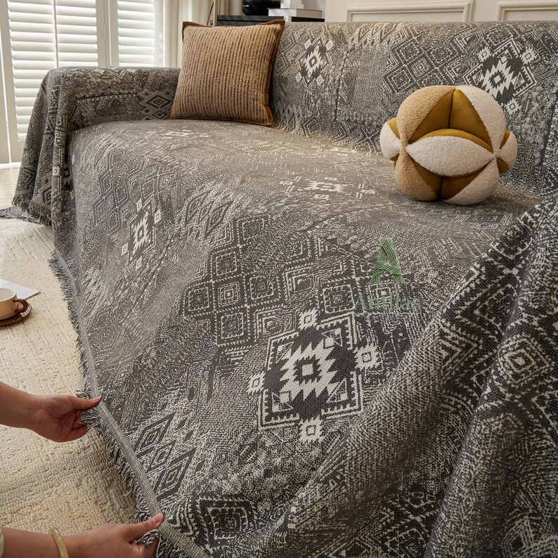 May include: A grey sofa cover with a geometric pattern and fringed trim. The cover is draped over a sofa, with a brown cushion and a decorative ball on top. The pattern includes white and dark grey shapes.