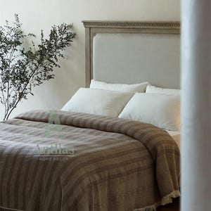 Pu&ograve; includere: Un letto con testiera grigio chiaro e cuscini bianchi. Una coperta a righe marroni con frange copre il letto. La coperta ha un aspetto strutturato. L'immagine mostra anche un paio di pantofole marroni sul pavimento.