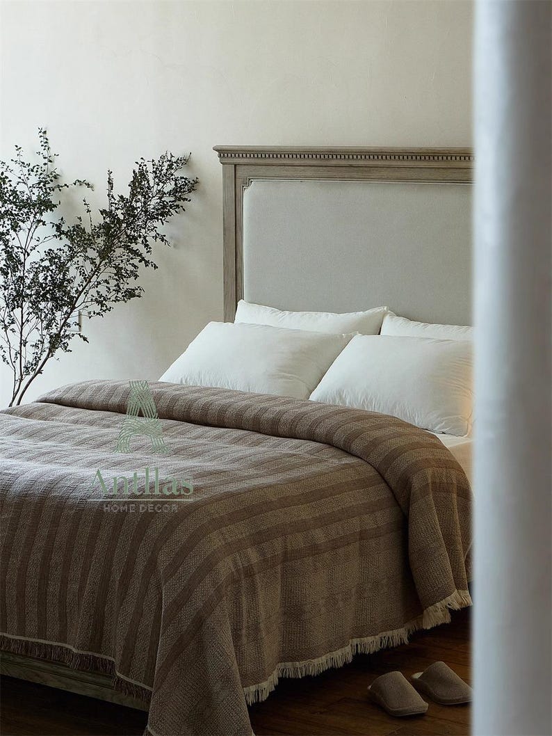 Pu&ograve; includere: Un letto con testiera grigio chiaro e cuscini bianchi. Una coperta a righe marroni con frange copre il letto. La coperta ha un aspetto strutturato. L'immagine mostra anche un paio di pantofole marroni sul pavimento.