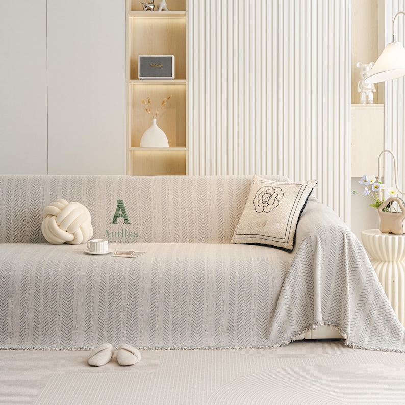 May include: A beige sofa with a patterned throw blanket, a decorative knot pillow, and a floral pillow. A small white teacup and saucer sit on the blanket. The word "Antilus" is printed on the blanket. A pair of slippers are on the floor.