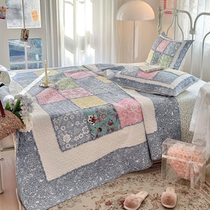 May include: A patchwork quilt and pillow set on a bed. The quilt has squares of pink, blue, yellow, and floral patterns. The bed is made up with a white bedspread and a decorative pillow. The room has a cozy, inviting atmosphere.