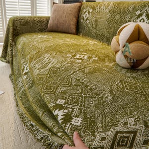 May include: A green sofa throw with a white geometric pattern is draped over a sofa. The throw has a fringed edge and is paired with a brown cushion. A decorative cushion with a tan and brown colour scheme is also on the sofa.