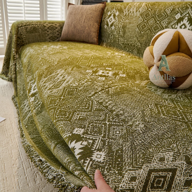 May include: A green sofa throw with a white geometric pattern is draped over a sofa. The throw has a fringed edge and is paired with a brown cushion. A decorative cushion with a tan and brown colour scheme is also on the sofa.