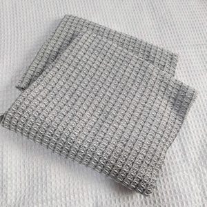 May include: Two folded gray waffle weave kitchen towels. The towels have a textured, geometric pattern and are resting on a white, textured surface. The towels are made of a soft, absorbent material.