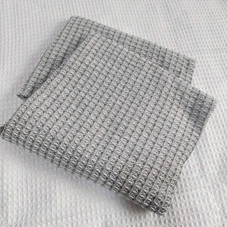 May include: Two folded gray waffle weave kitchen towels. The towels have a textured, geometric pattern and are resting on a white, textured surface. The towels are made of a soft, absorbent material.