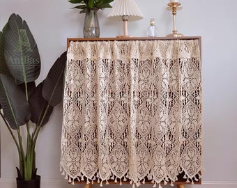 May include: A wooden cabinet with a cream-colored lace curtain. The curtain features a floral pattern and tassels along the bottom edge. A lamp, candle holder, and decorative items sit on top of the cabinet. A large potted plant is on the left.