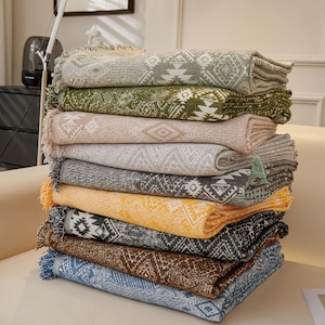 May include: A pile of ten folded blankets in shades of grey, green, beige, yellow, black, brown, and blue. Each blanket has a geometric design and fringed edges. The blankets are stacked on top of each other.