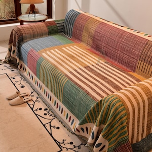 May include: A colorful sofa cover with a geometric pattern. The cover features stripes and blocks of red, green, blue, and tan. The edges are fringed. The sofa cover is draped over a sofa.