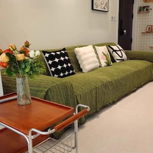 May include: A living room scene featuring a green sofa with decorative pillows. The pillows have various designs, including black and white polka dots, fluffy white, and floral patterns. A wooden serving cart with a metal frame and a vase of flowers are in the foreground. The text "Real customer scene" is at the bottom.