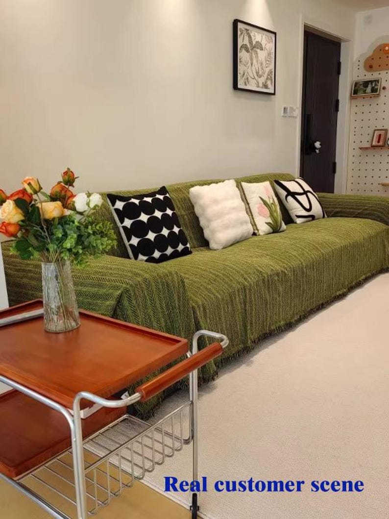 May include: A living room scene featuring a green sofa with decorative pillows. The pillows have various designs, including black and white polka dots, fluffy white, and floral patterns. A wooden serving cart with a metal frame and a vase of flowers are in the foreground. The text "Real customer scene" is at the bottom.