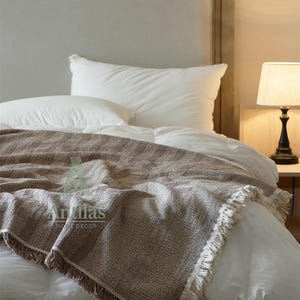 Pu&ograve; includere: Una coperta marrone intrecciata con frange &egrave; drappeggiata su un letto con biancheria da letto bianca. La coperta ha un aspetto strutturato e porta il testo "Antillas HOME DECOR". Una lampada con paralume beige &egrave; su un comodino.