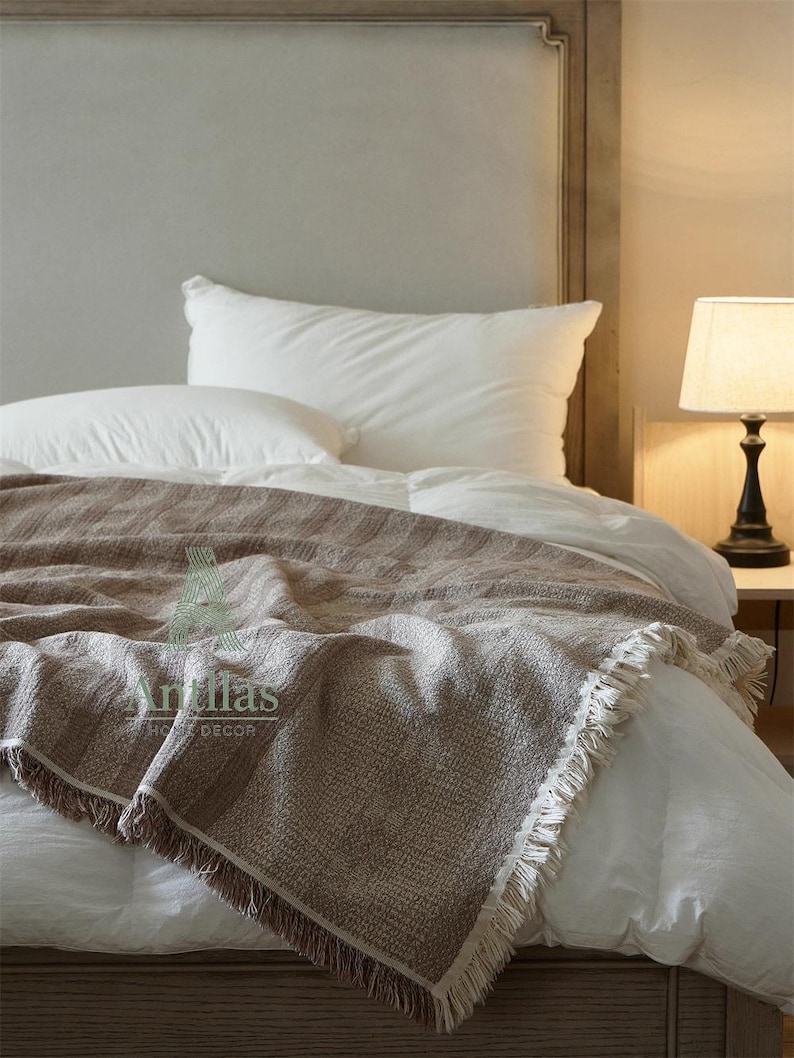 Pu&ograve; includere: Una coperta marrone intrecciata con frange &egrave; drappeggiata su un letto con biancheria da letto bianca. La coperta ha un aspetto strutturato e porta il testo "Antillas HOME DECOR". Una lampada con paralume beige &egrave; su un comodino.