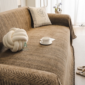 May include: A brown sofa with a patterned throw blanket and decorative pillows. A white knot-shaped pillow and a pillow with a floral design are visible. A white teacup and saucer sit on the sofa. The sofa is in a well-lit room.
