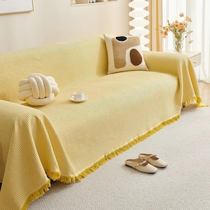 May include: A yellow waffle-weave sofa cover with a fringed edge. A white knot pillow and a decorative pillow with brown and black shapes rest on the sofa. A pair of beige slippers sits on the rug.