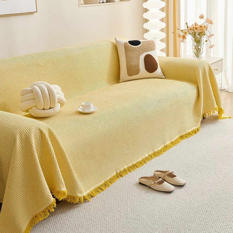May include: A yellow waffle-weave sofa cover with a fringed edge. A white knot pillow and a decorative pillow with brown and black shapes rest on the sofa. A pair of beige slippers sits on the rug.