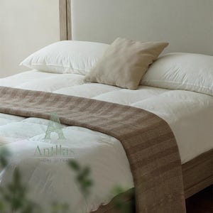 Pu&ograve; includere: Un letto con biancheria da letto bianca, due cuscini bianchi e una coperta marrone. Un cuscino quadrato beige poggia sui cuscini bianchi. La struttura del letto &egrave; in legno chiaro. Il logo "Antillas HOME DECOR" &egrave; visibile.