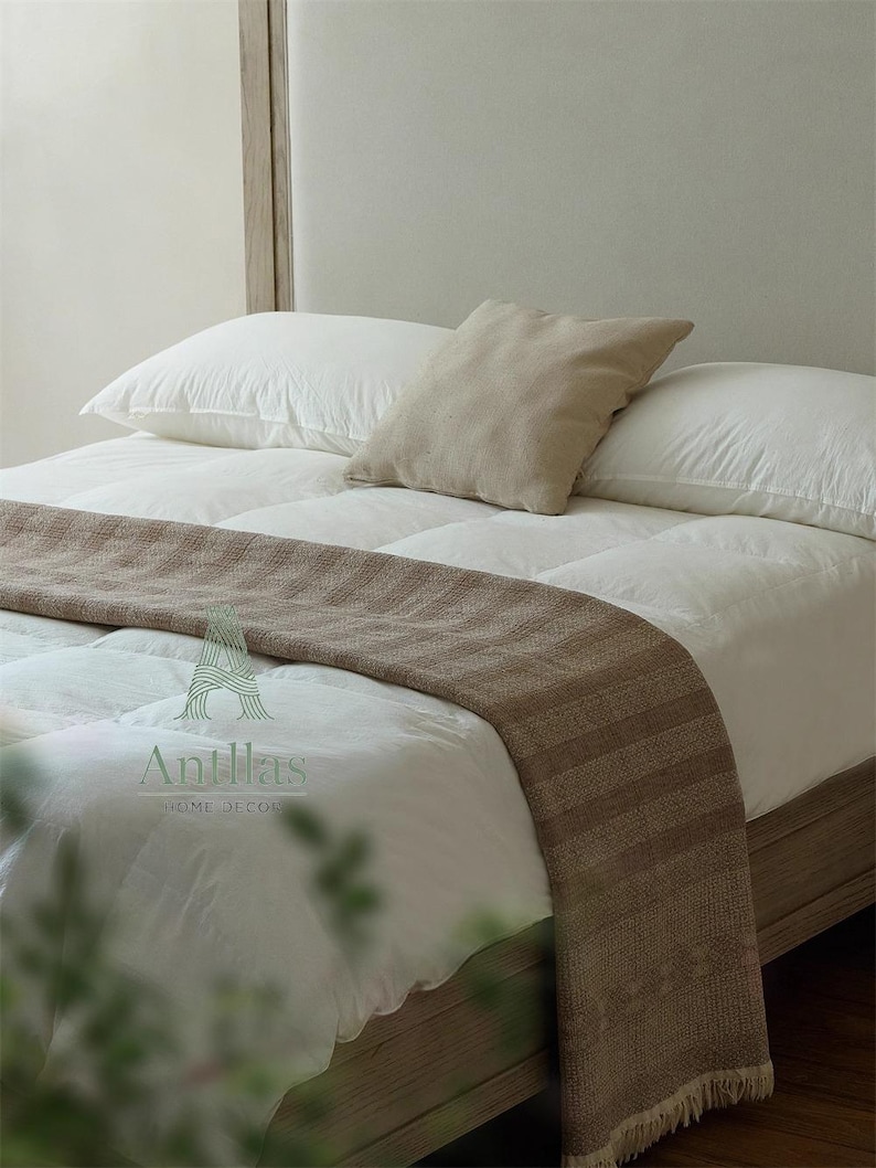 Pu&ograve; includere: Un letto con biancheria da letto bianca, due cuscini bianchi e una coperta marrone. Un cuscino quadrato beige poggia sui cuscini bianchi. La struttura del letto &egrave; in legno chiaro. Il logo "Antillas HOME DECOR" &egrave; visibile.