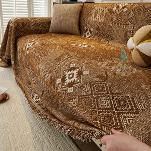 May include: A brown and white patterned sofa cover with a geometric design. The cover is draped over a sofa, with a matching cushion and a decorative ball visible. The cover has a fringed edge and a textured appearance.