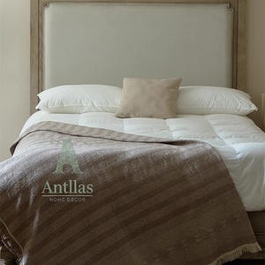 Pu&ograve; includere: Un letto con testiera colorata chiara, biancheria da letto bianca e una coperta a righe marroni. Un piccolo cuscino beige poggia sopra i cuscini. La coperta ha un bordo con frange e il testo "Antllas HOME DECOR".