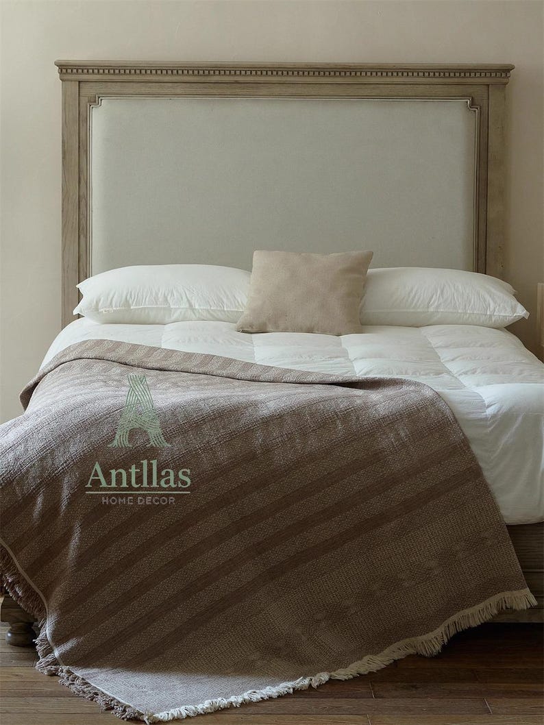 Pu&ograve; includere: Un letto con testiera colorata chiara, biancheria da letto bianca e una coperta a righe marroni. Un piccolo cuscino beige poggia sopra i cuscini. La coperta ha un bordo con frange e il testo "Antllas HOME DECOR".