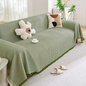 May include: A green and white checkered sofa cover with a fringe trim, draped over a sofa. Decorative pillows and a teacup sit on the sofa. White slippers are on the floor.