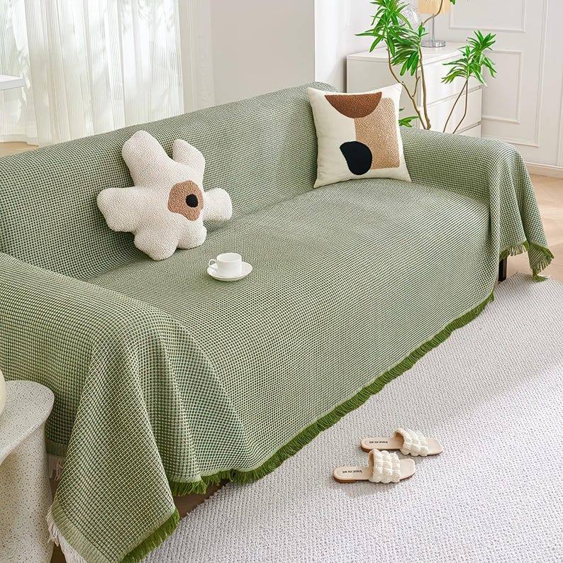 May include: A green and white checkered sofa cover with a fringe trim, draped over a sofa. Decorative pillows and a teacup sit on the sofa. White slippers are on the floor.