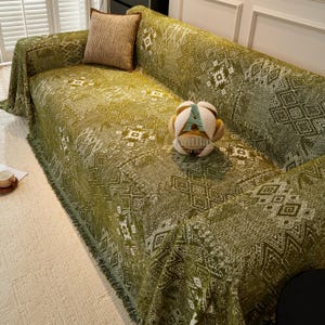 May include: A green sofa cover with a geometric pattern and fringed trim. A decorative cushion and a soft toy are on the sofa. The cover is draped over the sofa, offering protection and style.
