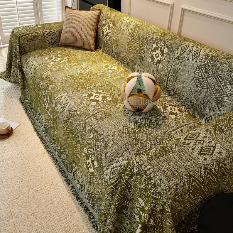 May include: A green sofa cover with a geometric pattern and fringed trim. A decorative cushion and a soft toy are on the sofa. The cover is draped over the sofa, offering protection and style.