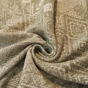 May include: Close-up of a textured fabric with a repeating geometric pattern in shades of olive green and off-white. The fabric is draped, showing folds and depth, with a woven appearance.
