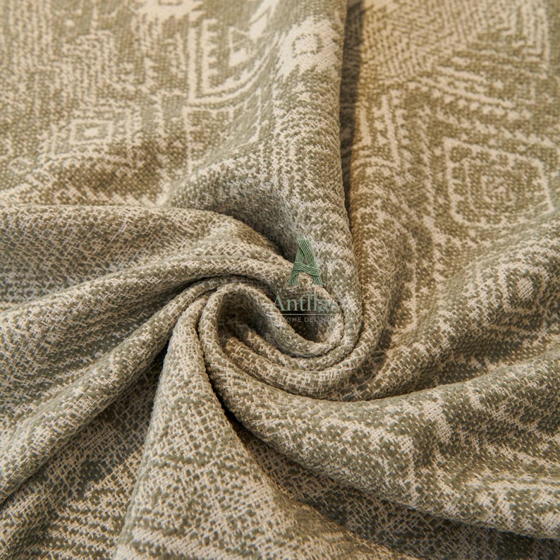 May include: Close-up of a textured fabric with a repeating geometric pattern in shades of olive green and off-white. The fabric is draped, showing folds and depth, with a woven appearance.