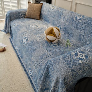May include: A blue and white patterned sofa cover with fringe trim. A brown square cushion and a geometric ball are on the sofa. The cover has a geometric design and is draped over the sofa.
