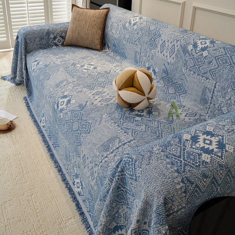 May include: A blue and white patterned sofa cover with fringe trim. A brown square cushion and a geometric ball are on the sofa. The cover has a geometric design and is draped over the sofa.