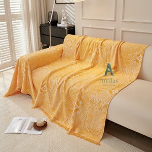 May include: A yellow sofa throw with a geometric pattern and fringed edges. The throw is draped over a white sofa. The text "Antllas HOME DECOR" is visible on the throw.