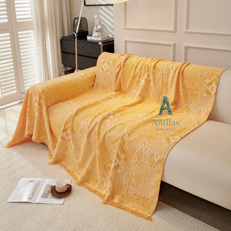 May include: A yellow sofa throw with a geometric pattern and fringed edges. The throw is draped over a white sofa. The text "Antllas HOME DECOR" is visible on the throw.