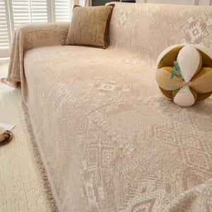 May include: Beige sofa cover with a geometric pattern and fringe trim. A matching pillow with a brown border rests on the sofa. A decorative, round, multi-textured pillow is also on the sofa. The sofa is in front of a window with white shutters.