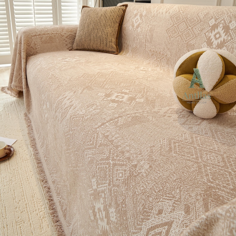 May include: Beige sofa cover with a geometric pattern and fringe trim. A matching pillow with a brown border rests on the sofa. A decorative, round, multi-textured pillow is also on the sofa. The sofa is in front of a window with white shutters.