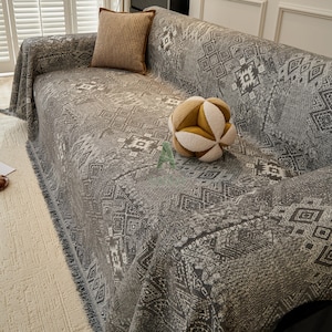 May include: A grey sofa cover with a geometric pattern, fringed edges, and a matching throw pillow. A decorative spherical pillow with yellow and white segments rests on the sofa. The sofa is in a well-lit room.