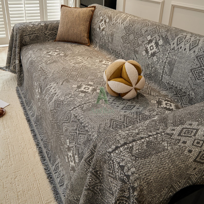 May include: A grey sofa cover with a geometric pattern, fringed edges, and a matching throw pillow. A decorative spherical pillow with yellow and white segments rests on the sofa. The sofa is in a well-lit room.