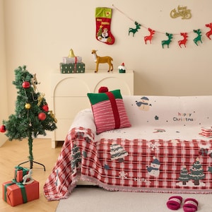 Puede incluir: Una escena navideña festiva con un árbol decorado, un sofá con una manta temática navideña y elementos decorativos. Un cojín rojo y verde con forma de regalo está en el sofá. Un calcetín navideño y una guirnalda cuelgan de la pared.